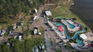 Aerial drone view of Fireside RV Resort showing the winding lazy river, swimming pools, RVs parked among tall pine trees, and the resort's lake
