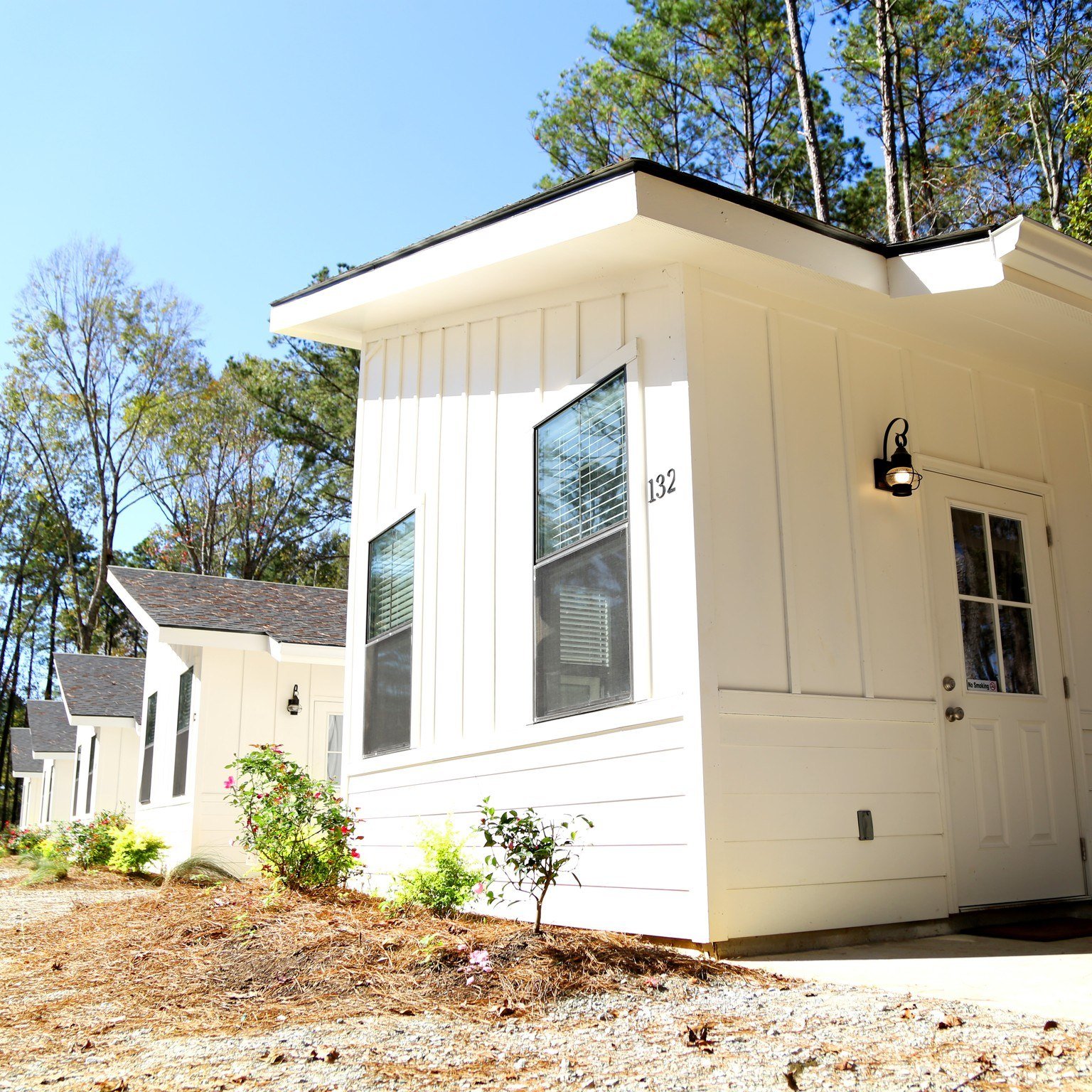 A modern white cabin with a covered entry, large windows, and a porch light, set among tall pine trees at Fireside RV Resort