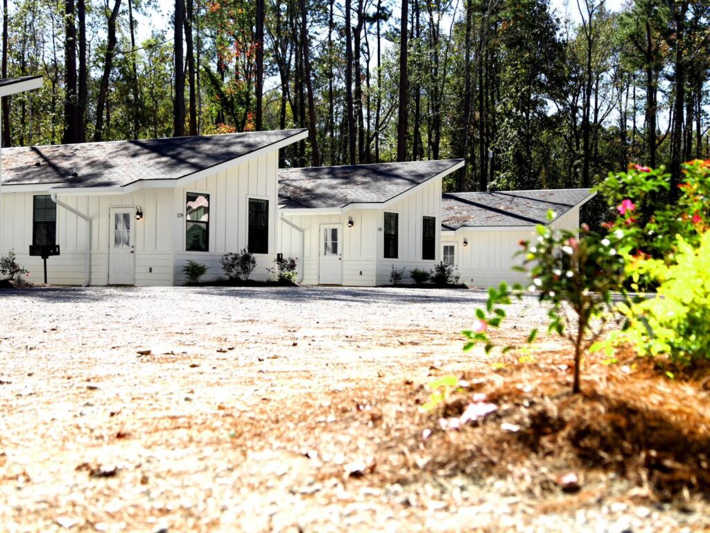 Row of modern white cabins with dark trim at Fireside RV Resort lined along a gravel drive backed by tall Louisiana pines on a sunny day