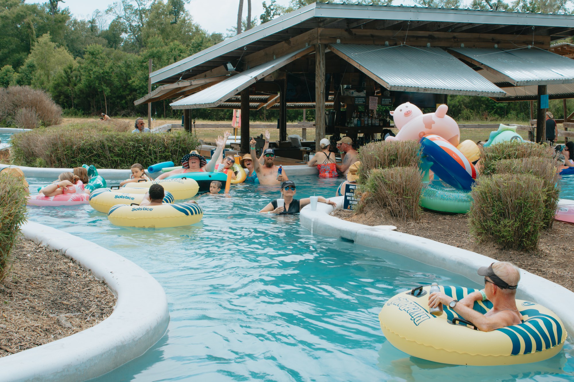 Guests floating on inflatable tubes along the lazy river at Fireside RV Resort, passing a covered swim-up bar on a sunny summer day