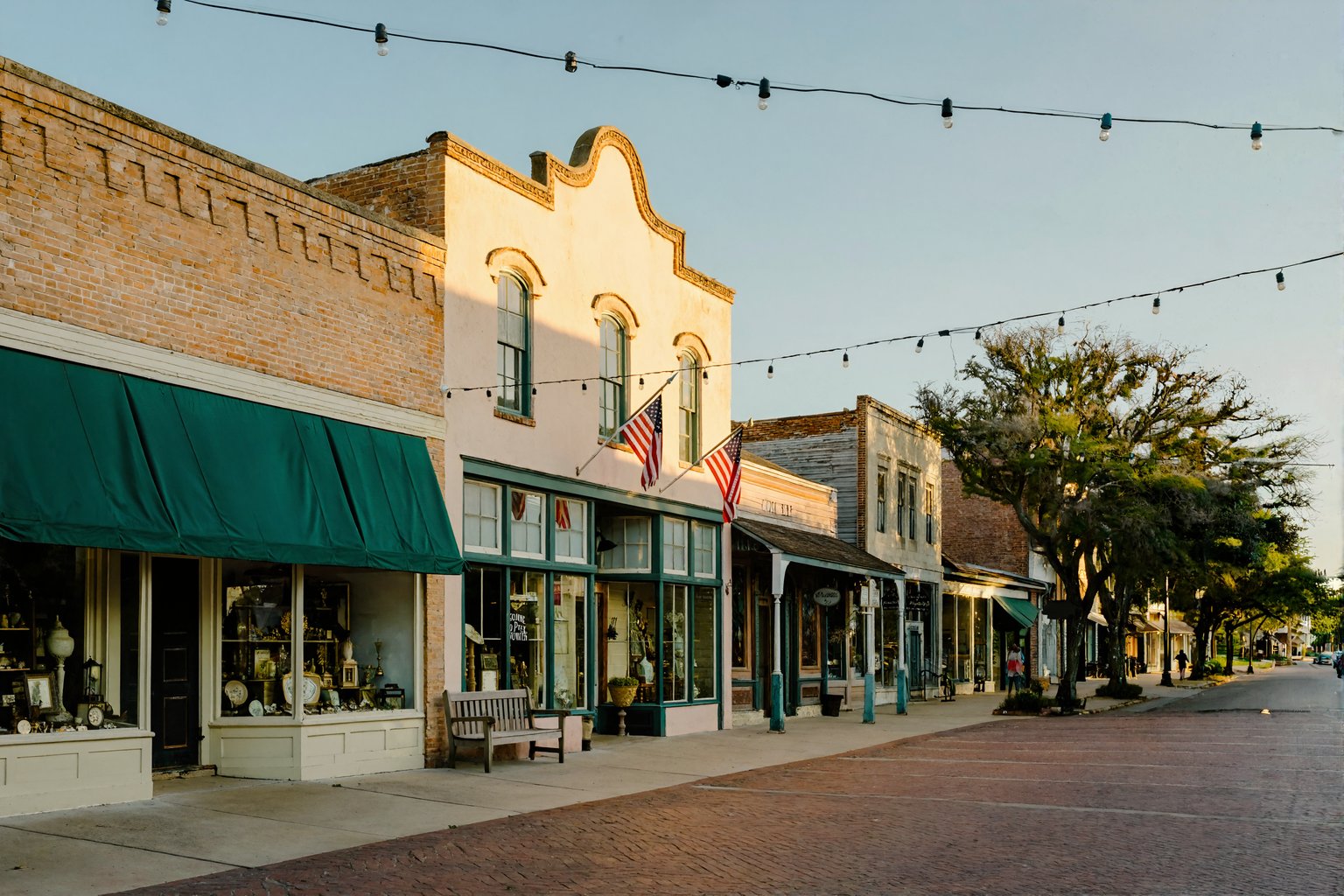 A quiet historic small-town main street near Ponchatoula at golden hour, lined with antique-shop storefronts under green awnings, American flags, and bistro string lights over a brick sidewalk